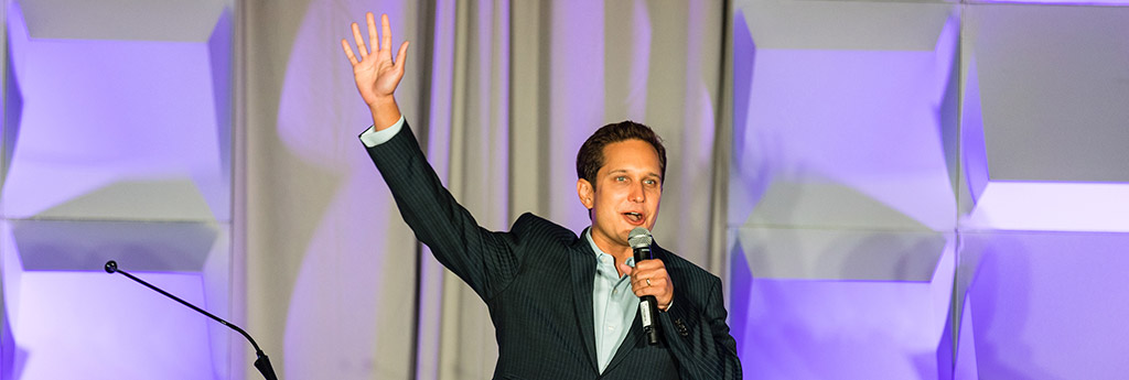 Jason Dorsey speaking in-person on a stage with his right hand waving and a purple lighted background